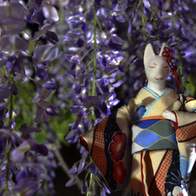Fox in a plum blossom kimono standing beneath illuminated purple wisteria flowers