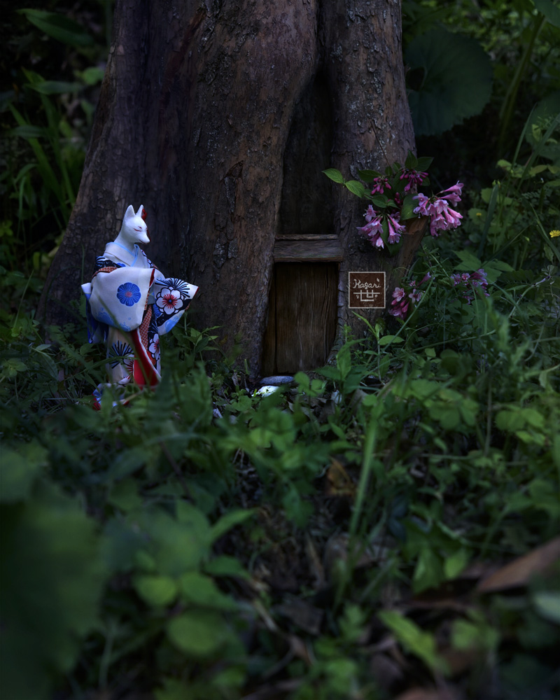 Fox standing before a small door in the hollow of a large tree root among early morning grass