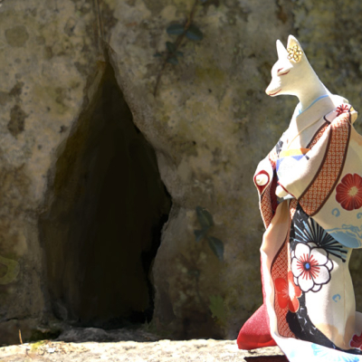 White kimono-wearing fox standing beside a shimenawa-wrapped garden stone with a cool dark hollow